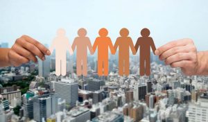 community, unity, population, race and humanity concept - multiracial couple hands holding chain of paper people pictogram over city background